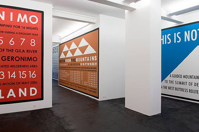 Sobre las paredes blancas de una galería están varios carteles a gran escala de color rojo, café y azul con información e iconos de montañas.