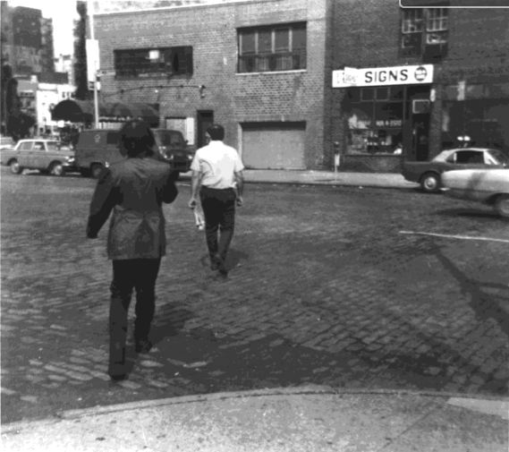  Fotografía en blanco y negro de una escena urbana en la que vemos de espaldas a Acconci y, a unos pasos delante de él, al hombre que persigue.
