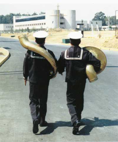 Fotografía a color en la que vemos de espaldas a dos personas uniformadas marchar juntas cargando dos mitades de una tuba grande. La campana sale bajo el brazo derecho de una mientras el cuello curvado envuelve la espalda de la otra.