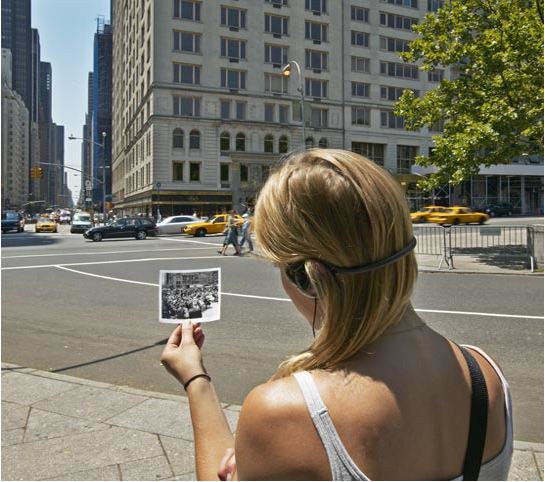 Fotografía a color de una mujer rubia de espaldas en una calle de la Ciudad de Nueva York. Tiene puestos audífonos y sostiene una fotografía en blanco y negro. 