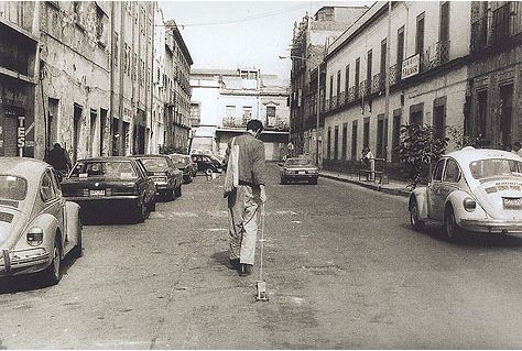 Fotografía en blanco y negro en la que vemos de espaldas al artista jalando un carrito cuadrado con forma de perro en medio de una calle urbana. 
