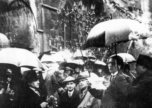  Fotografía en blanco y negro de personas reunidas alrededor de un árbol. Muchas de ellas llevan paraguas y casi todas miran hacia un hombre con hojas de papel en las manos.