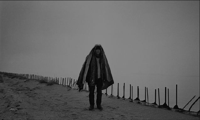 Fotografía en blanco y negro de un hombre cubierto por una cobija que camina por un paisaje con arena y neblina.