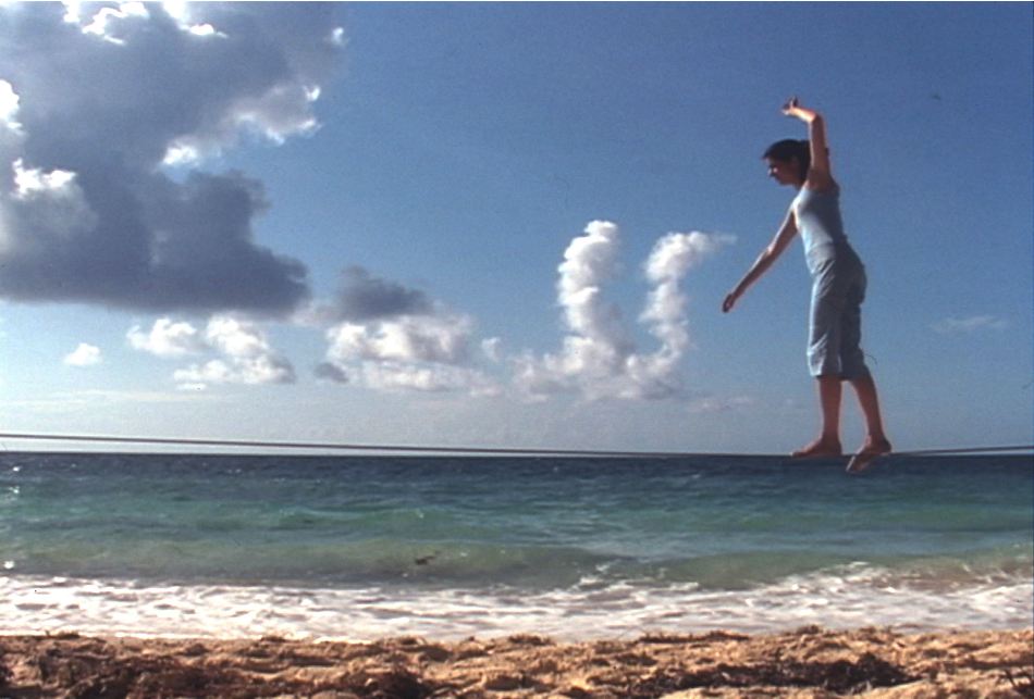 Fotografía a color en la que una cuerda floja se extiende sobre el mar y crea una línea paralela a la del horizonte. Vestida de azul, Antoni mantiene el equilibrio mientras camina sobre la cuerda. 