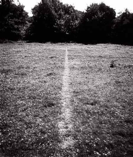 Fotografía en blanco y negro de un campo. En medio del césped reluce una línea recta donde el pasto ha sido aplastado. No se puede distinguir dónde comienza o termina.
