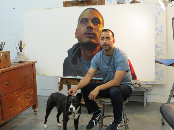 Un hombre de cabello corto, barba incipiente y tez morena acaricia a un perro color blanco y negro frente a una pintura que retrata a otro hombre.