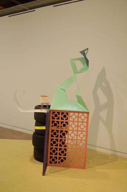 Jessica Stockholder, Assist #3: A Chord, 2015. Painted metal, ratchet clamp with yellow webbing, felt (if needed), some kind of found sup- port to clamp the work to. 74 x 90 x 48 inches. 