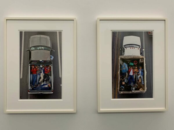 Sobre una pared blanca, dos fotografías a color están expuestas una junto a la otra. Ambas retratan desde arriba a personas acostadas en la parte de atrás de camionetas de carga.