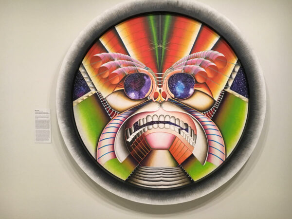 Colorida pintura en forma de círculo con borde blanco en el que una imagen superpuesta sobre formas geométricas recuerda a una máscara de dioses aztecas con gafas de sol que reflejan el cosmos.