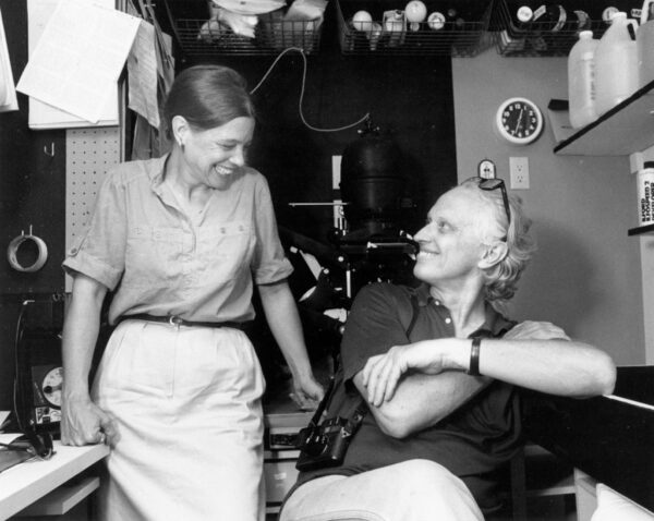 A black and white photograph of a seated White man and standing White woman. The figures are side by side, looking and smiling at each other. They are located inside a small photographic darkroom with equipment above and around them.