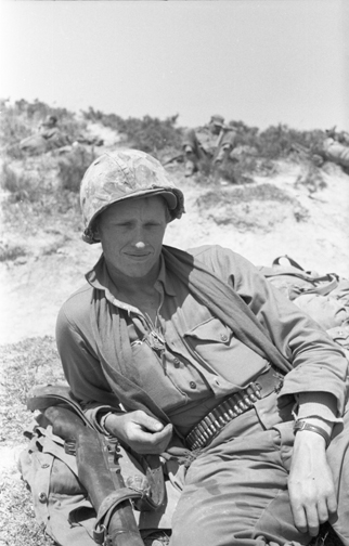 A low contrast black and white image of a young United States Marine. The figure sits outdoors in a relaxed pose looking straight into the camera. 