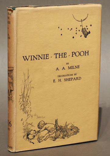 Photograph of the book "Winnie the Pooh" by A. A. Milne. The book appears slim and the book cover is tan with dark writing. The bottom left corner has an illustration of a young boy and several rabbits attempting to pull a bear from an underground hole. The top right has an illustration of the bear floating in the sky, holding onto a ballon.
