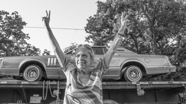 A black and white photograph of Ann Harithas standing in front of a car. She stands in the center of the image, looking into the distance with a smile on her face. Both arms are raised above her head in a "v" shape. Both hands are holding up the gesture for peace.