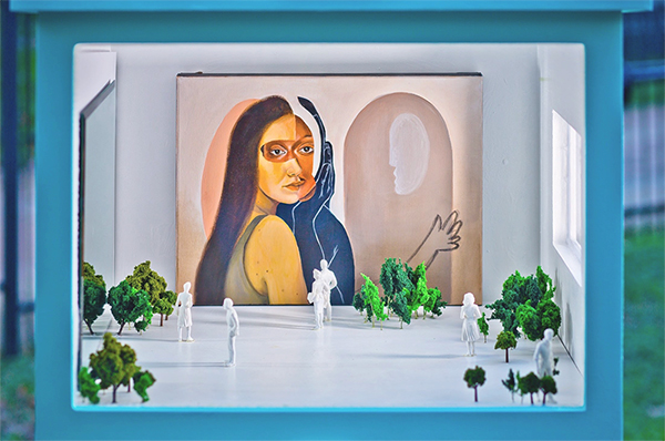 A photograph of a small-scale installation inside a little gallery on a stand, similar in size to a Free Little Library. The installation includes a small painting of an abstracted female figure looking into a mirror as well as small trees and small white figures standing in front of the painting. The artwork is by Margo Lunsford.
