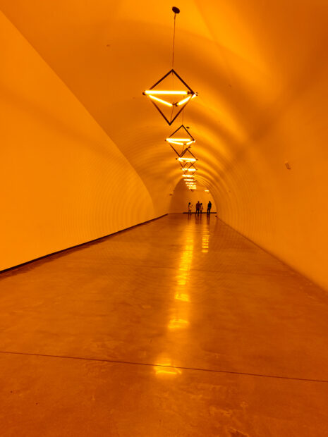 A vertical photograph of the inside of a tunnel walkway with multiple hanging lights, each in the shape of a triangle. The lights glow a bright yellow and immerse the tunnel in yellow light. Installation by Ólafur Elíasson.
