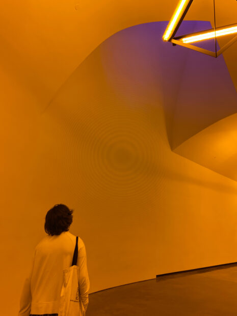 A visitor looks up at a blue-violet light that interrupts the bright yellow lighting inside a tunnel walkway. Installation by Ólafur Elíasson.