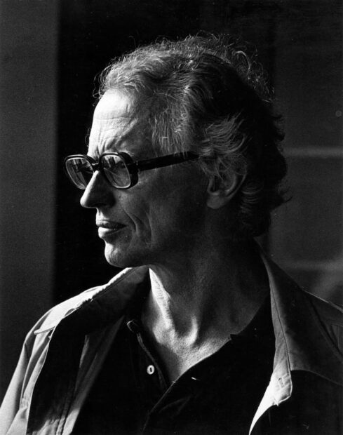 A high contrast black and white photographic portrait of an older man with head turned revealing his profile. The man wears dark rimmed glasses and looks intently into the distance.