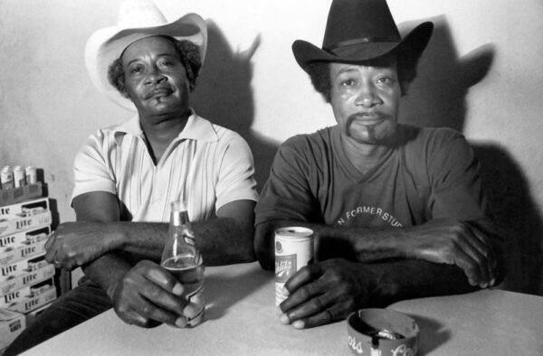 A black and white photograph of two Black men sitting side by side at a table with drinks in hand. The men look straight into the camera and appear to be at the end of a work day. Both wear cowboy hats and short sleeved shirts.
