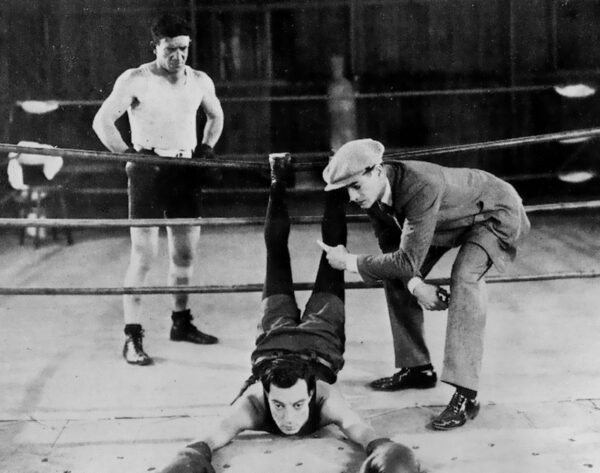 Film still from Buster Keaton's "Battling Buster." A black and white image of a boxer laying on the floor of the boxing ring with arms stretched out in front of him and legs in the air with his feet resting on the top rope. A man in a suit leans over the boxer. Another boxer can be seen watching in the background.
