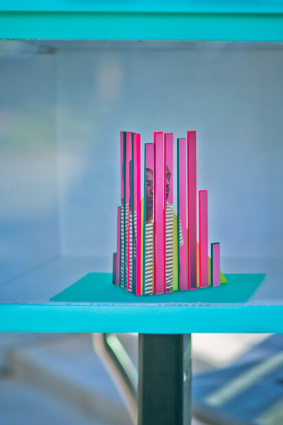 A close-up photograph of a small-scale sculpture inside a little gallery on a stand, similar in size to a Free Little Library. The sculpture is made up of vertical strips some of which are bright pink and others depict fragments of a photograph of a person. The artwork is by Karen Navarro.