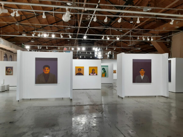 A photograph of an art exhibition. The photo features many mid-size portrait paintings hung on walls.