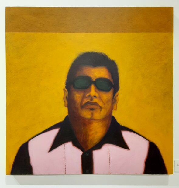 A portrait of a figure wearing sunglasses and a pink and black shirt. The figure stands in front of a yellow background.