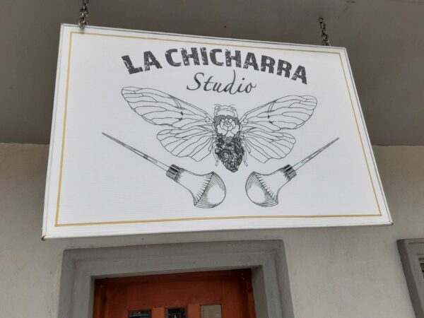 Un letrero blanco con dibujos simples de una chicharra con alas abiertas y dos goteras de tinta dice en letras negras “La Chicharra Studio”.