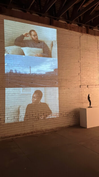 A photograph of a video projection on the wall of a gallery. The projection is three videos, stacked, and features one landscape image of a city in the middle, and a figure each on the top and bottom.