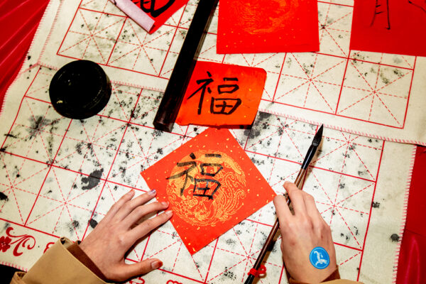 A photograph of two hands writing in a calligraphy style. One hand holds a brush, the other hand holds the red paper in place on the table top.