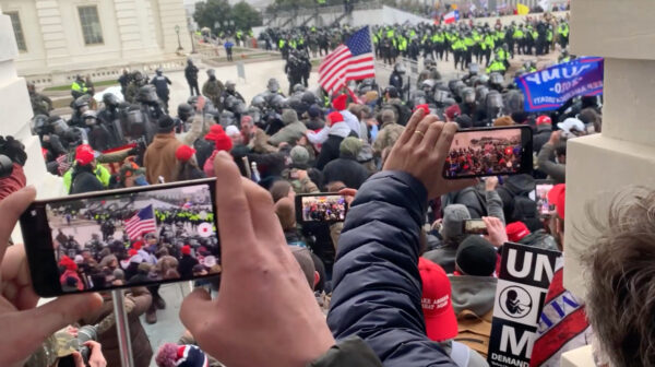 A photograph of a large crowd of people standing on the steps of the US Capitol building. Many people are holding up their phones, and many are also wearing Donald Trump-related clothing. 