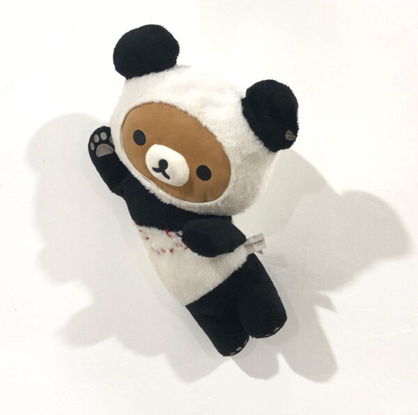 Statue of a small stuffed animal.  The animal has a brown face and appears to be wearing a panda bear costume.  The chest of a stuffed animal is embroidered with red thread, creating two "AT" shapes where the pectoral muscles would be.  Artwork by Angela Faz