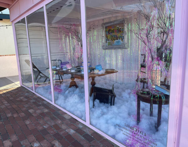 A color photograph of a shipping container painted pink with the one side of the container replaced with windows. Through the windows you can see an exhibition inside the space that includes a large dining table with cake sculptures, white stuffing covering the floor, and iridescent streamers covering the background. A framed map of the United States with text that reads, "Eat It & Weep," is hung on the back wall. Artwork by Sarah Ayala and Deryk Poynor.