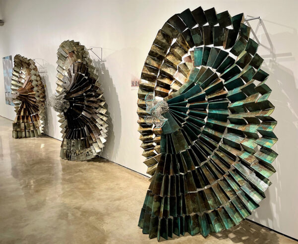 A photograph of three large-scale sculptures by Naomi Wanjiku. Each sculpture is a spiral of accordion folded sheet metal and resembles a flowing skirt.
