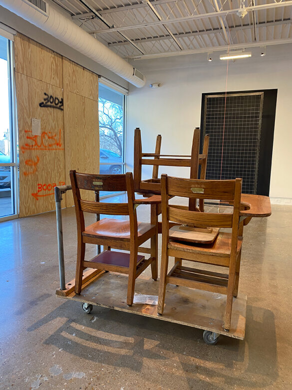 An installation of works by Benjamin Loftis including three wooden chairs stacked on a dolly, a window covered with plywood, and a large grid structure standing in front of a black painted background.