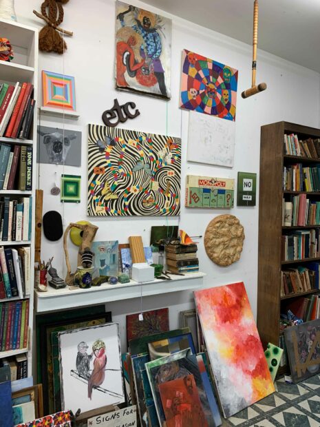 A photograph of a crowded store, featuring books, paintings, and other objects on shelves and the store's floor.