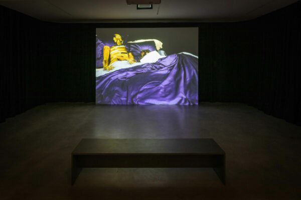 Installation photo of a video still of two figures in a bed with purple satin linens. One figure appears to be made of metal.