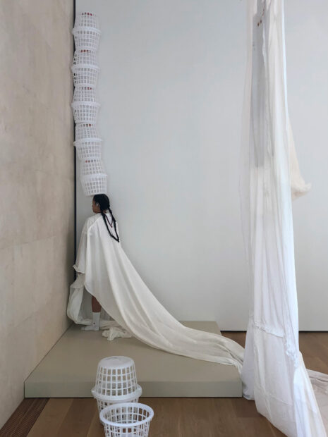 A performance artwork by artist Christian Cruz. Cruz is standing in the corner of an art gallery. She is wearing white and balancing a stack of laundry baskets on her head.