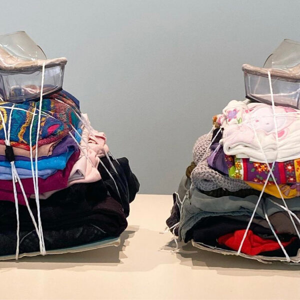 Two stacks of laundry, each sitting on a table and comprised of various garments. Both stacks are topped with a shoe.