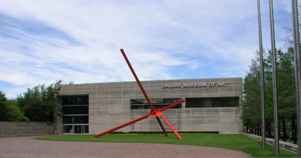 A photo of the Dallas Museum of Art. The building is gray, and there is a large, red metal sculpture sitting in front of the building.