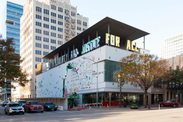 A photograph of the outside of The Contemporary Austin. There is a green lift placed in front of the large street facing exterior wall. A mural of artwork by Daniel Johnston is in process.