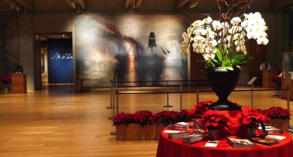 Entrance to the Exhibition "Turner's Modern World" on view at the Kimbell Museum. The image shows a table with a vase of flowers in the foreground, and a large scale, gestural painting of a nautical scene in the background