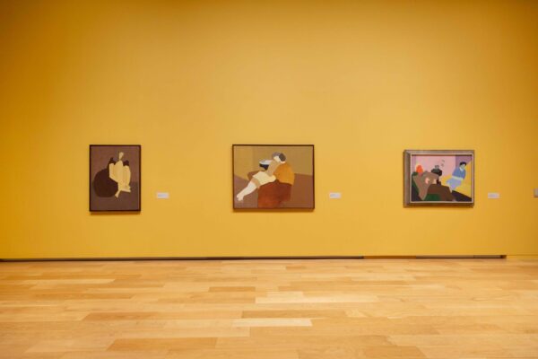 En una pared amarilla del museo cuelgan tres cuadros en los que sujetos humanos dominan el espacio pictórico. Los tres comparten una paleta de tonos terrosos, aunque en el último también resaltan los colores brillantes de azul, verde y naranja.