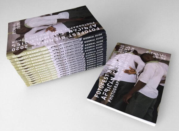 A color photograph of a stack of books. On the left is a stack of thirteen thin books. The text on each book spine reads, "FotoFest Biennial 2020 African Cosmologies Biennial Guide." On the right is the one copy of the same book. The cover shows a photograph of three young Black boys with their backs turned to the camera. The text on the cover reads, "FotoFest Biennial 2020 African Cosmologies Photography, Time, and the Other. Biennial Guide." 