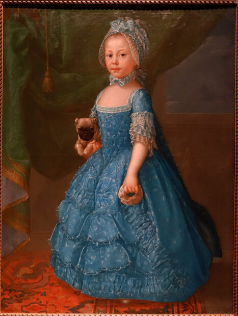 A painting of a women. She is standing, wearing a blue dress, and holding a dog in her right hand. Her surroundings are very opulent. 
