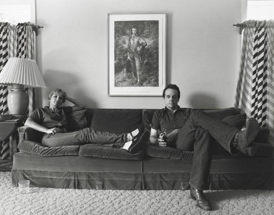 A black and white photograph of Linda and Ed Blackburn sitting on a dark couch inside a home. Both sit in a relaxed style with legs outstretched and crossed. Photograph by Gay Block.