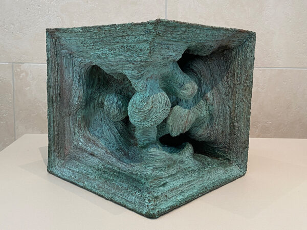 A photograph of a sculpture made by Harry Bertoia. The sculpture is generally in the shape of a cube with two sides removed revealing an interior that is filled with more organic shapes. It was created by welding layered pieces of bronze and the layers are evident in the final work. The metal has oxidized and appears to be a light blue-green color.