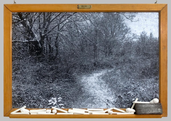 A painting using acrylic on a vintage chalkboard to create the illusion that the painting is made with chalk. The image in the painting is a very realistic depiction of a wooded area in winter with little foliage. A snowy path meanders through the trees. The work also includes sticks of chalk, plaster acorns, and an eraser set on the ledge of the chalkboard. Artwork by Helen Altman. 
