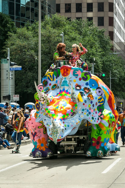 A sculpture of a brightly painted triceratops made to completely cover a vehicle. The car is participating in a parade and people are standing at the top of the dinosaur.