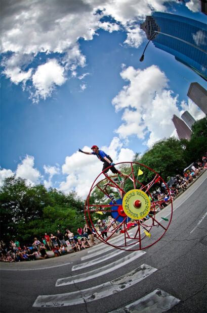 A man stands atop a large metal wheel as it rolls down the street in the Houston Car Art Parade.