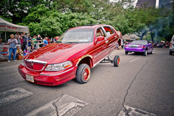 A candy red painted lowrider with gold pin striping participates in the Houston Art Car Parade. It's back end is lifted in the air using ha hydraulic system. 
